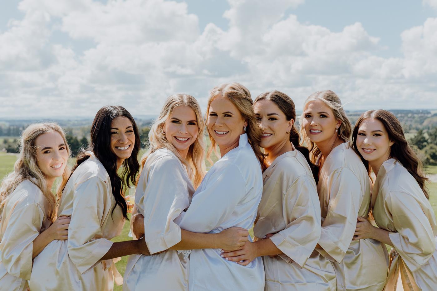 Bridesmaids in matching robes laughing together in a field under a bright sky