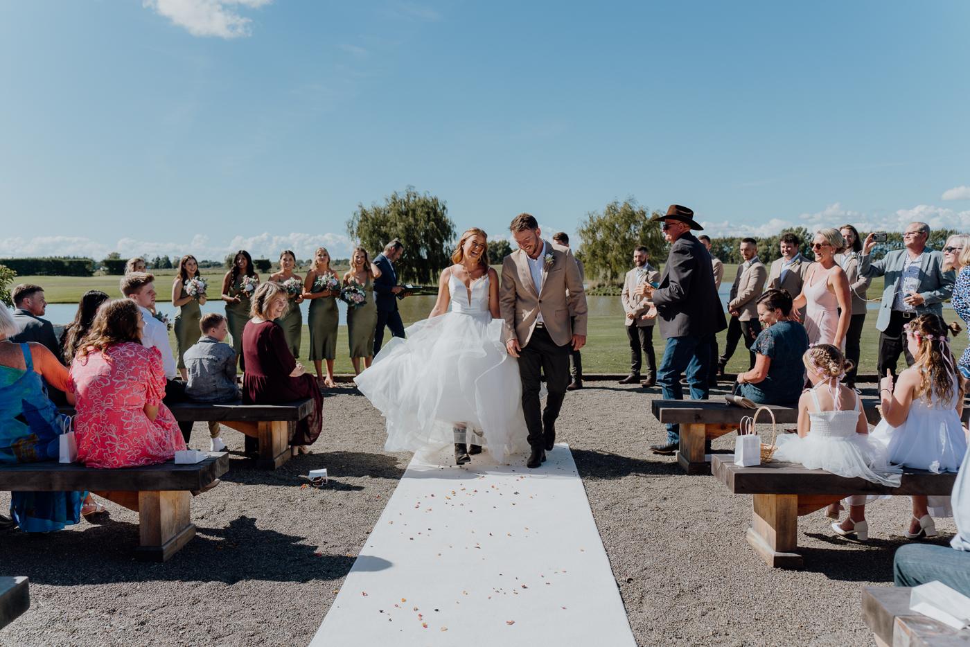 A bride and groom walking down a white carpet aisle between guests on benches