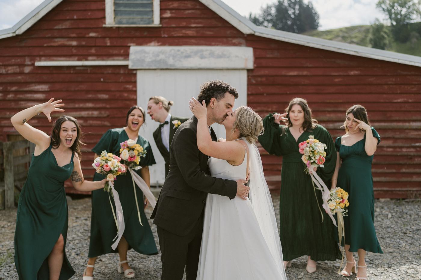 Bridal party in green dresses laughing and kissing outside a red barn