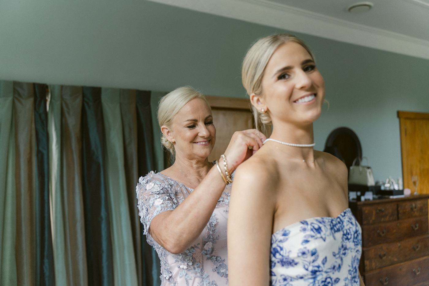A mother fastening a necklace on a laughing bride