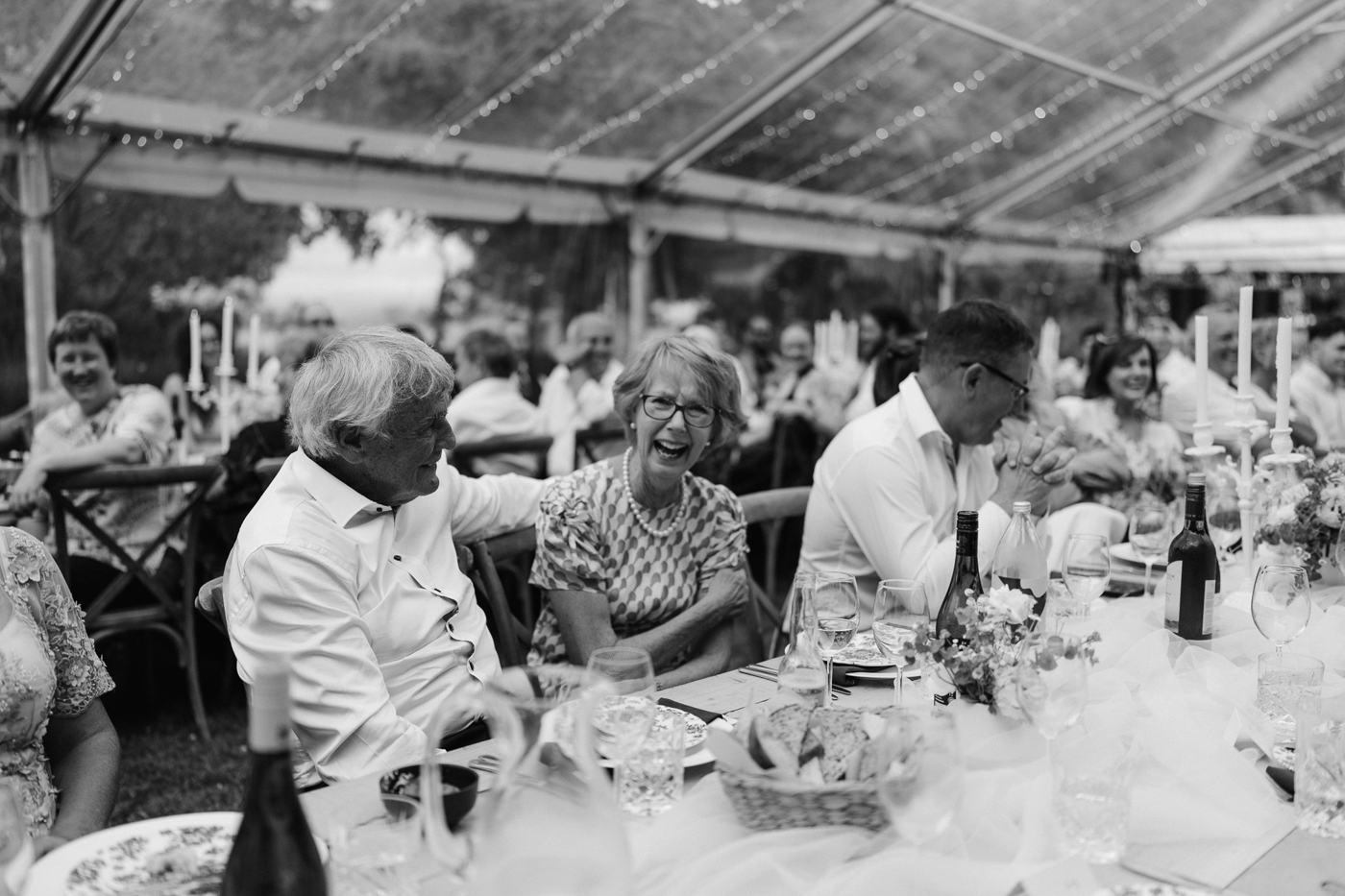 Guests laughing together at a long table in a marquee, in black and white