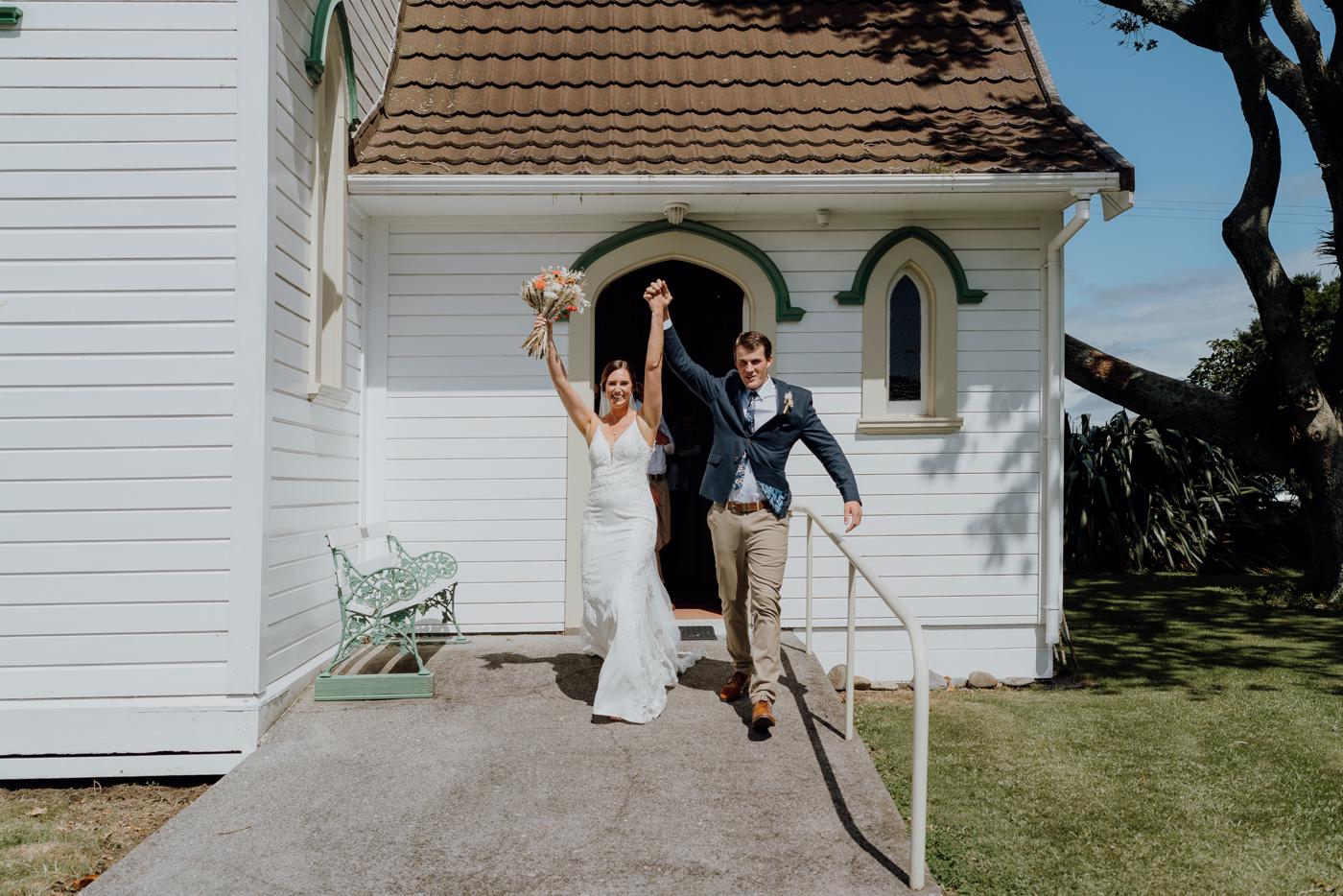 A bride and groom cheering as they leave a small white chapel