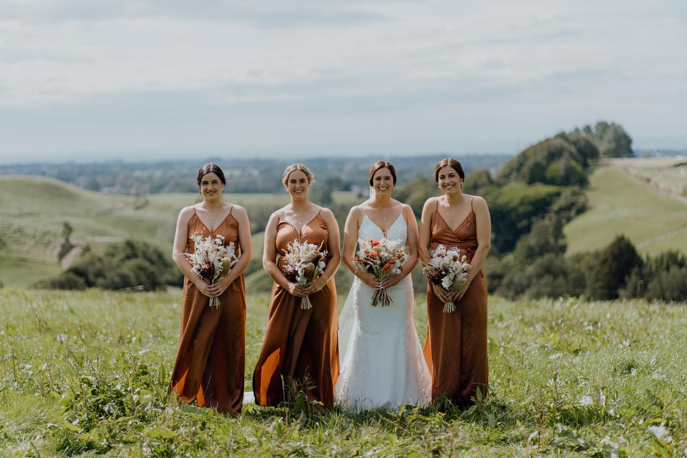 A bride flanked by bridesmaids in rust-coloured dresses on a hilltop