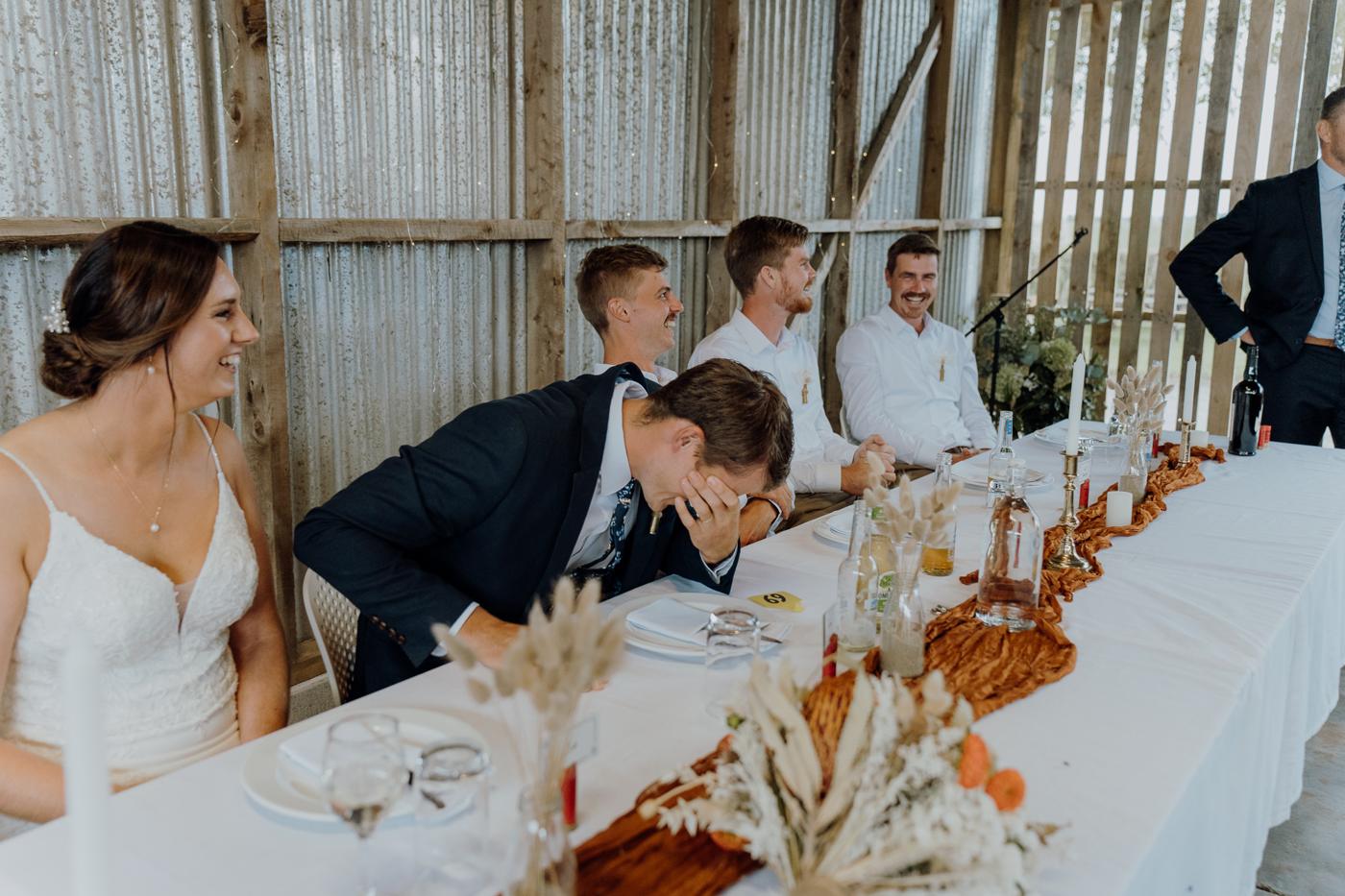 A groom leaning in to kiss his bride at a long table in a barn
