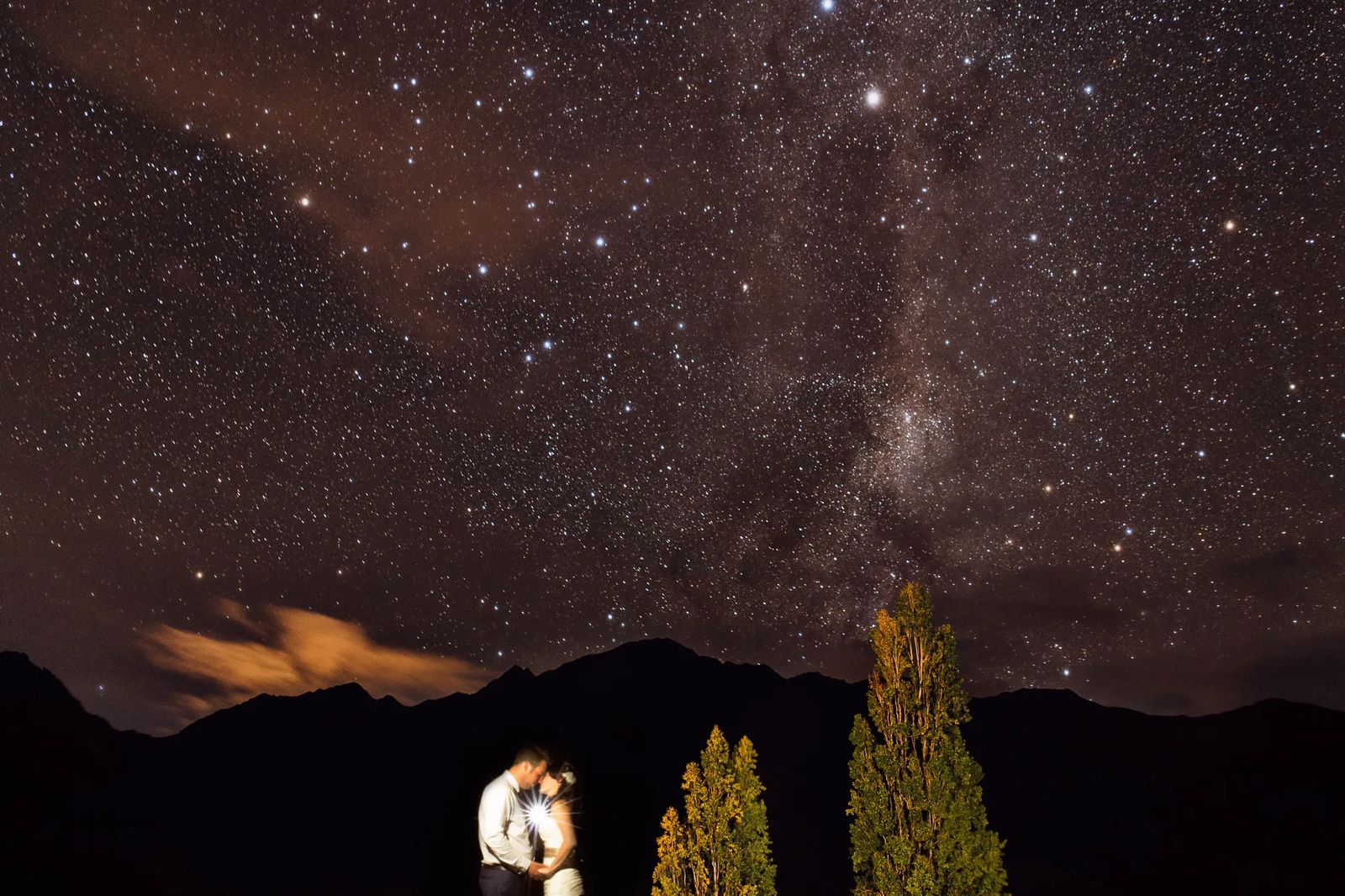 A couple embracing under the Milky Way with mountains behind