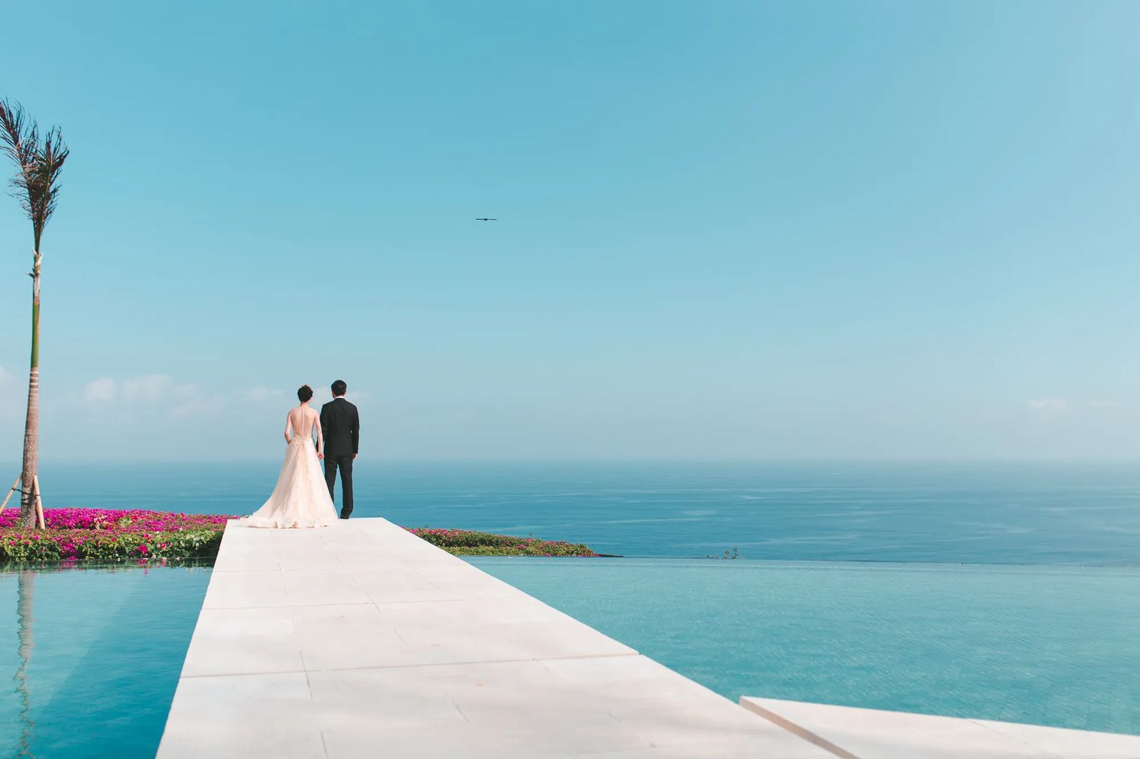 A couple standing at the end of a white jetty above a turquoise pool and sea