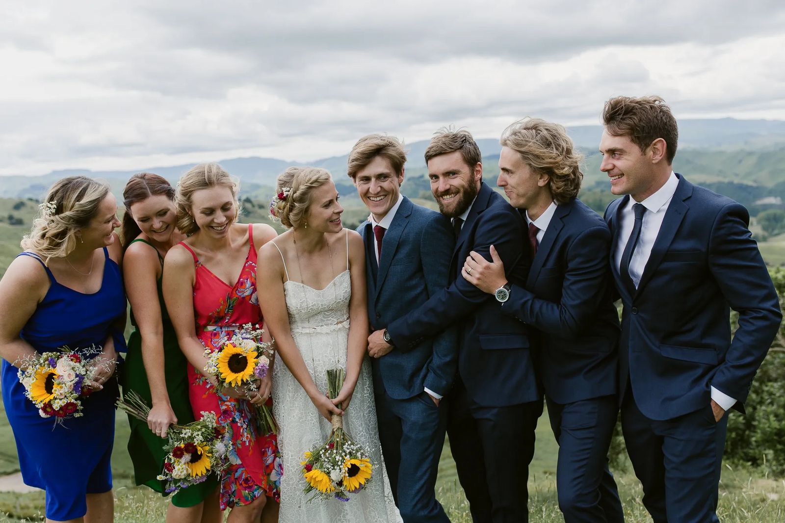A bridal party laughing together on a hillside, holding sunflower bouquets